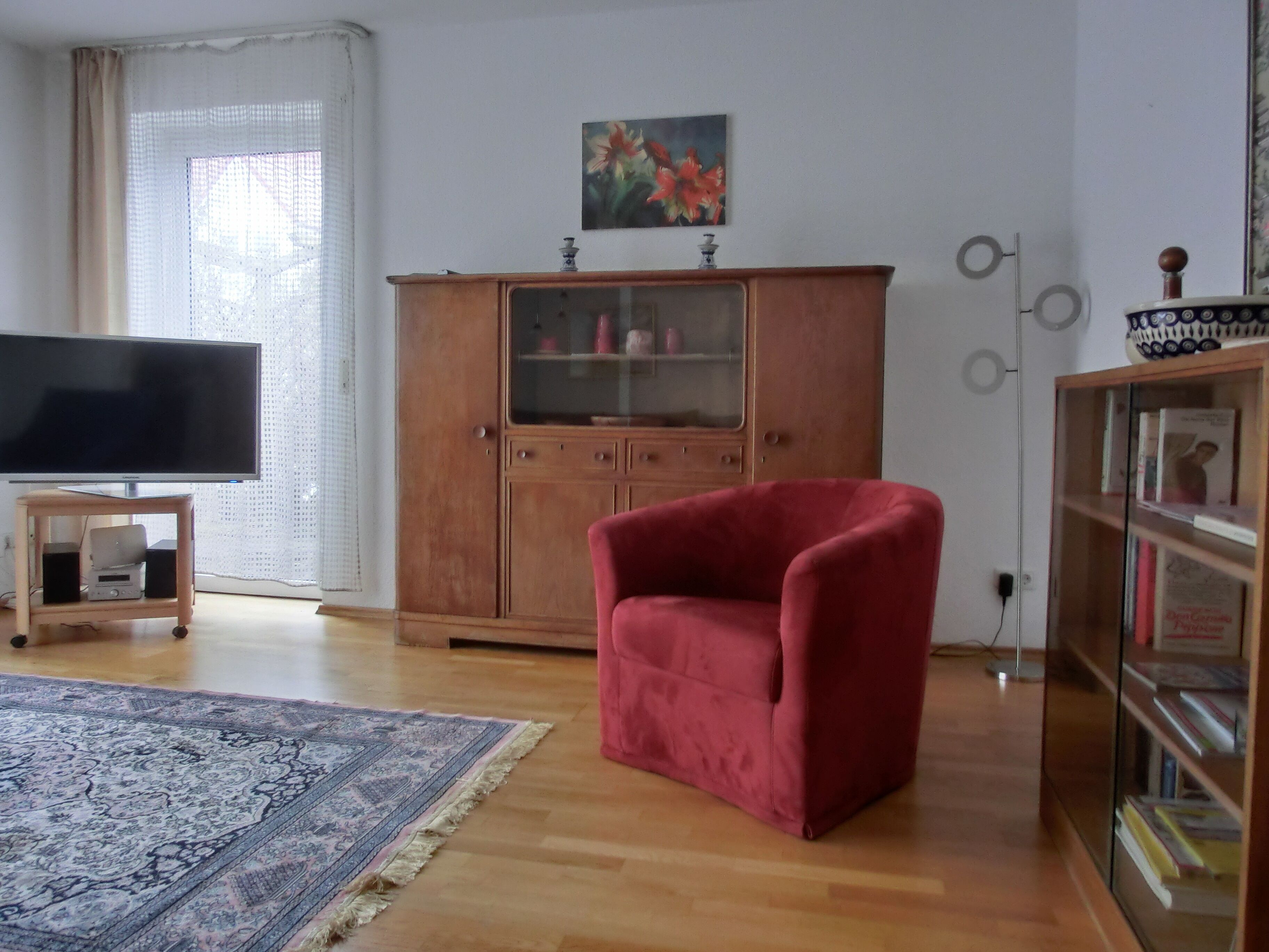 Ett vardagsrum med en platt-TV, en röd fåtölj, ett träskåp med glasdörrar och en mönstrad matta.
