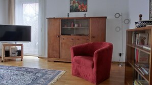 A living room with a flat-screen TV, a red armchair, a wooden cabinet with glass doors, and a patterned rug. - 3-zi.-fewo in Angenehmer und Ruhiger Lage im Erdgeschoss für 6 Pers (Graal-Müritz)