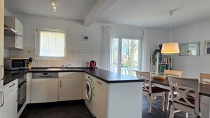 Eine moderne Küche mit weißen Schränken, einer schwarzen Arbeitsplatte und einem integrierten Geschirrspüler. Es gibt ein Fenster mit Vorhängen, eine Spüle und einen Essbereich mit Tisch und Stühlen.