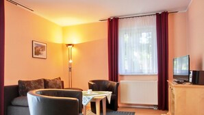 A living room with a sofa, two armchairs, a small table, a floor lamp, a radiator, a television, and a window with curtains. - Strandnahe 3-zimmer-fewo im EG für 4 Personen (Graal-Müritz)