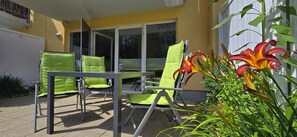 An outdoor patio with green cushioned chairs and a table, surrounded by plants and a yellow building. - 2-room apartment for 4 people on the first floor with Wi-Fi   (Graal-Müritz)