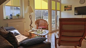 Ein gemütliches Wohnzimmer mit einer Couch, einem Korbsessel, einem Tisch mit Lampe sowie einem Fernseher an der Wand.