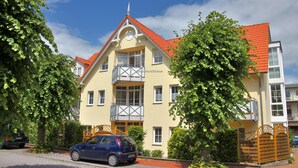 Ein zweistöckiges Gebäude mit rotem Ziegeldach und einem Balkon im zweiten Stock. Vor dem Haus ist ein Auto geparkt.