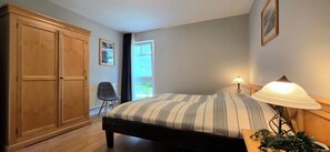 A bedroom with a bed, a nightstand, a lamp, a chair, a wardrobe, and a window.