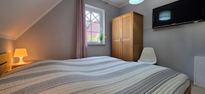 A bedroom with a bed, a nightstand, a lamp, a wooden wardrobe, a chair, a television, and a window with curtains. - Doppelhaushälfte mit ca. 95 m² Wohnfläche für 8 Personen (Graal-Müritz)