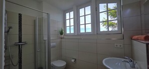 Un baño con cabina de ducha de vidrio, inodoro, lavamanos y ventana con vistas a árboles.