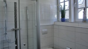 Un baño moderno con ducha de vidrio, paredes cubiertas de azulejos blancos, una ventana con vista a un edificio y una planta en el alféizar.
