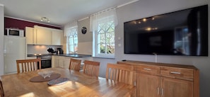 A modern kitchen with white cabinets, a dining table with chairs, a large flat-screen TV, and a window with curtains. - Doppelhaushälfte mit ca. 95 m² Wohnfläche für 8 Personen (Graal-Müritz)