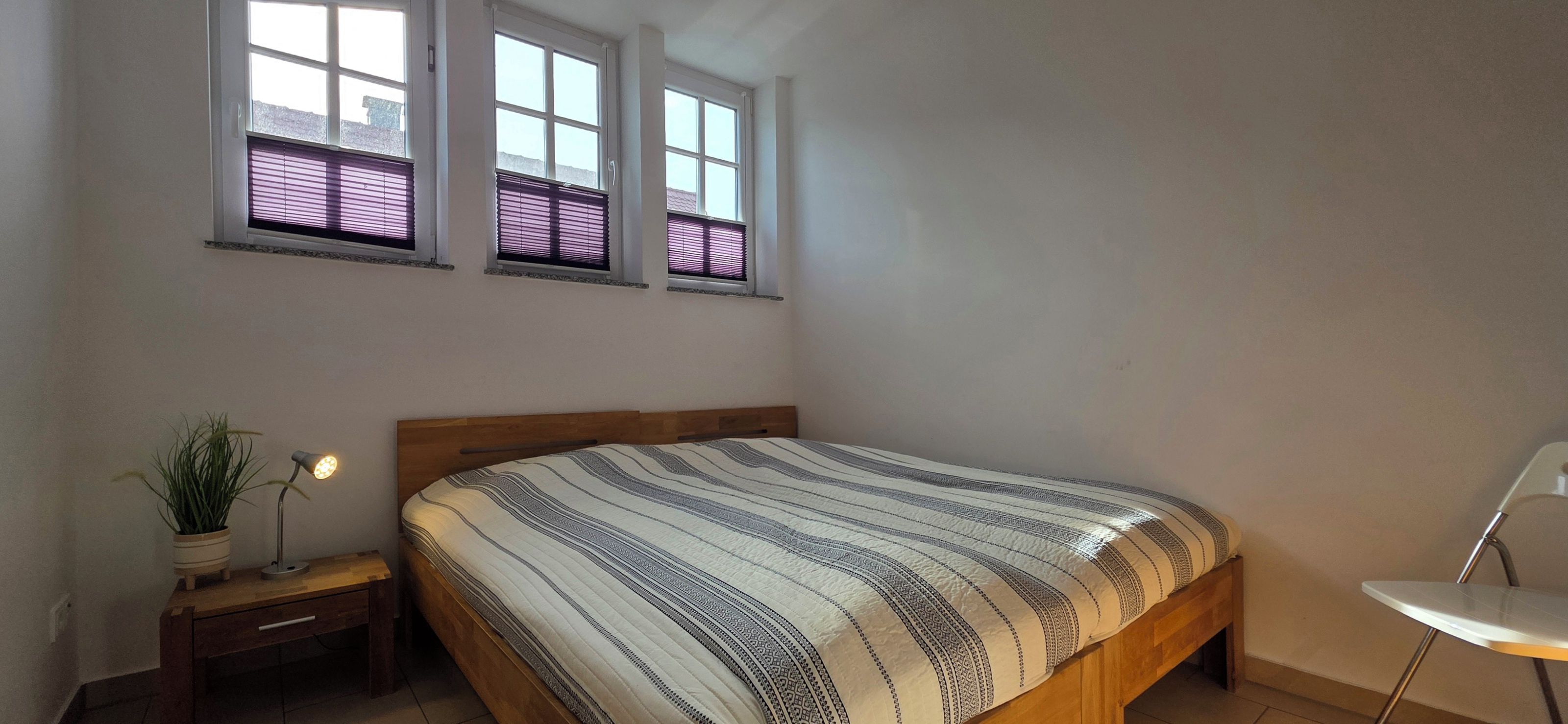 Ein Schlafzimmer mit einem Bett, einem Nachttisch, einer Topfpflanze und zwei Fenstern mit Jalousien.