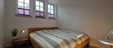 Ein Schlafzimmer mit einem Bett, einem Nachttisch, einer Topfpflanze und zwei Fenstern mit Jalousien.