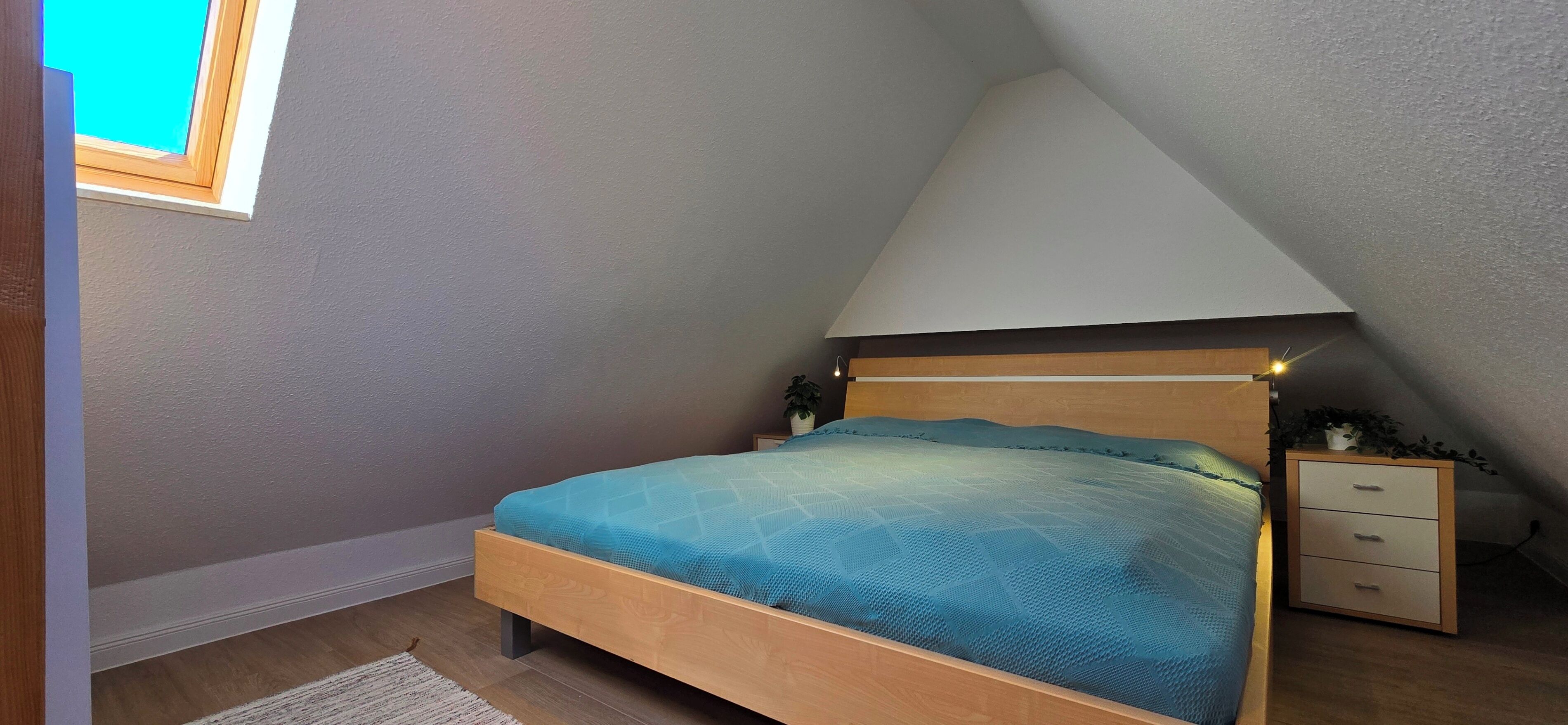 A bedroom with a sloped ceiling, a wooden bed frame, a blue bedspread, a bedside table with a plant, and a small drawer unit.