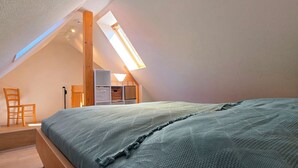 Un dormitorio con techo inclinado, tragaluz, cama con manta texturizada, una silla de madera y un pequeño escritorio con una cesta.