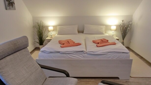 A double bed with white linens and two orange towels, a grey armchair, and a bedside table with a lamp. - 2- Zi.-fewo für 2 Personen mit Balkon und Wlan (Graal-Müritz)