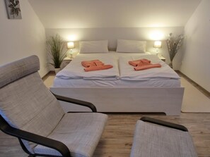 A double bed with white linens and two orange towels, a grey armchair, and a bedside table with a lamp.
