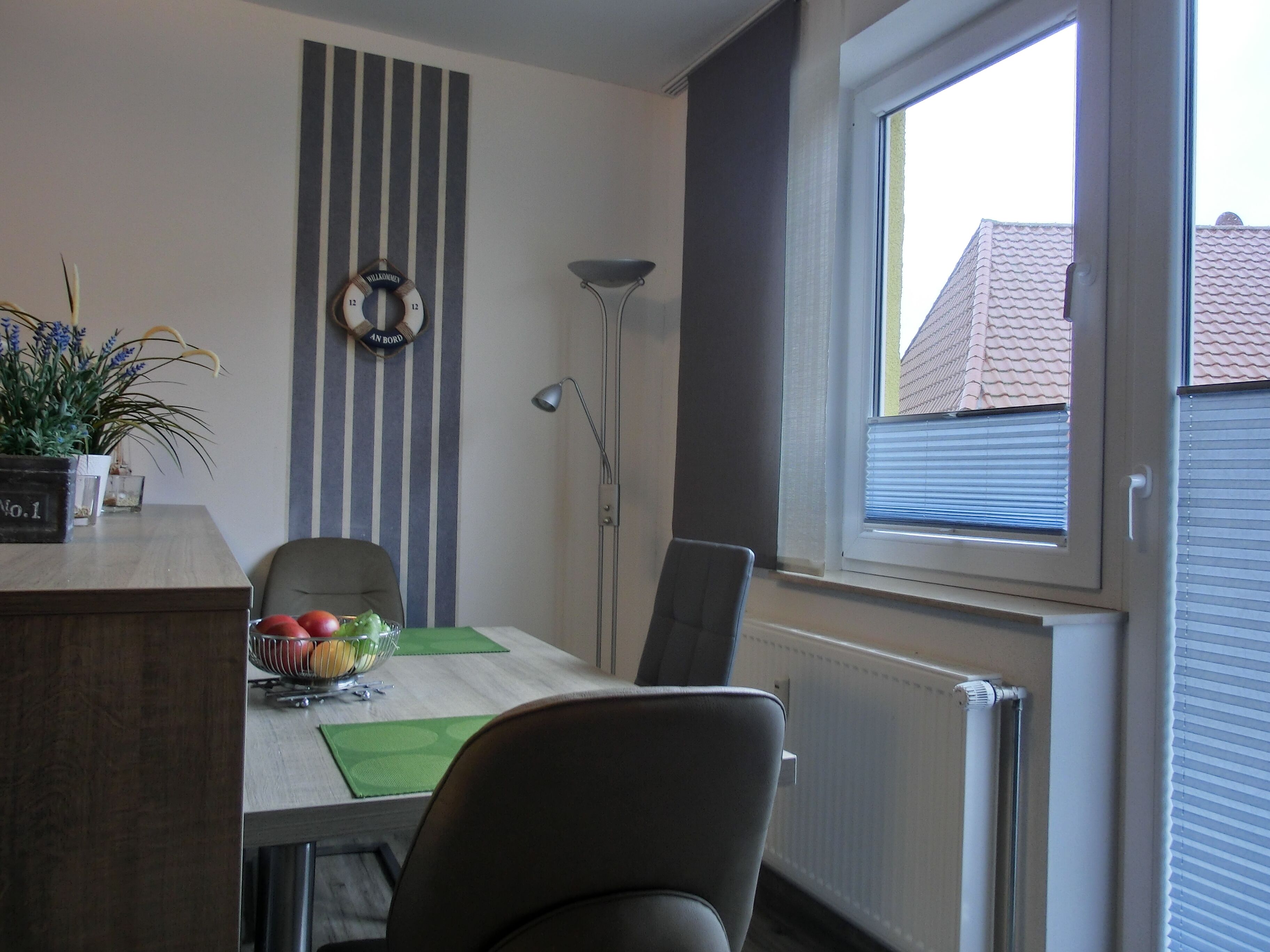 Un coin repas comprenant une table, des chaises, un saladier de fruits, une plante en pot et une horloge murale.