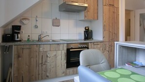 A compact kitchen with wooden cabinets, a built-in oven, a coffee maker, and a small dining area with a chair and table. - 2- Zi.-fewo für 2 Personen mit Balkon und Wlan (Graal-Müritz)