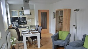 Ein modernes Wohnzimmer mit grauem Sofa und Sessel, einem Esstisch für zwei Personen und einer Küche im Hintergrund.