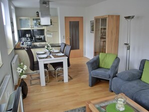 Una sala de estar moderna con un sofá y un sillón grises, una mesa de comedor para dos personas, y una cocina al fondo.
