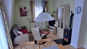 A living room with a sofa, a dining table with a blue plate, a TV, a cabinet, and a clock on the wall. - 3-zimmer-appartement im 1. OG mit Wlan (Graal-Müritz)