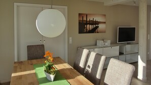 Ein Essbereich mit einem Holztisch, einer grünen Tischdecke und einem Blumentopf mit orangefarbenen Blüten. Es gibt vier gepolsterte Stühle, eine weiße Kommode mit einem Flachbildfernseher und ein Bild an der Wand.
