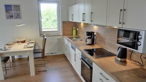 Une cuisine moderne avec des armoires blanches, un comptoir en bois, un four encastré, un four à micro-ondes et une machine à café. Une fenêtre laisse entrer la lumière naturelle et offre une vue sur la verdure environnante.