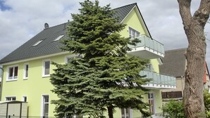 Une maison à deux étages, avec une façade verte, un toit gris et un balcon doté de rampes en verre.