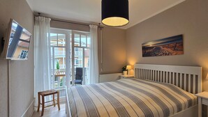 Un dormitorio con cama, mesita de noche, lámpara, ventana con cortinas y vistas a balcones, además de un cuadro en la pared.