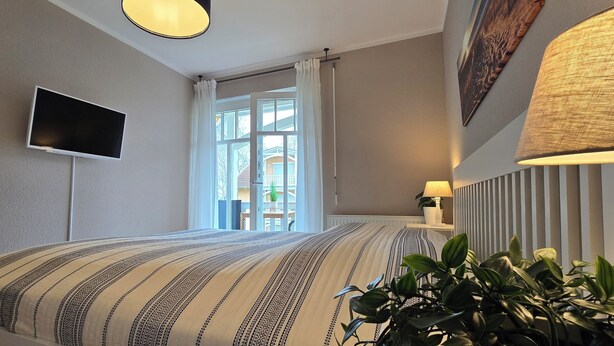 Una habitación de hotel con una cama grande, un televisor de pantalla plana montado en la pared, una ventana con cortinas y una planta en maceta.