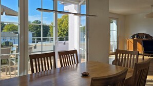 Un'area pranzo con un tavolo e sedie in legno, ampie finestre che offrono vista su un balcone e alberi, e un armadietto in legno con oggetti decorativi.