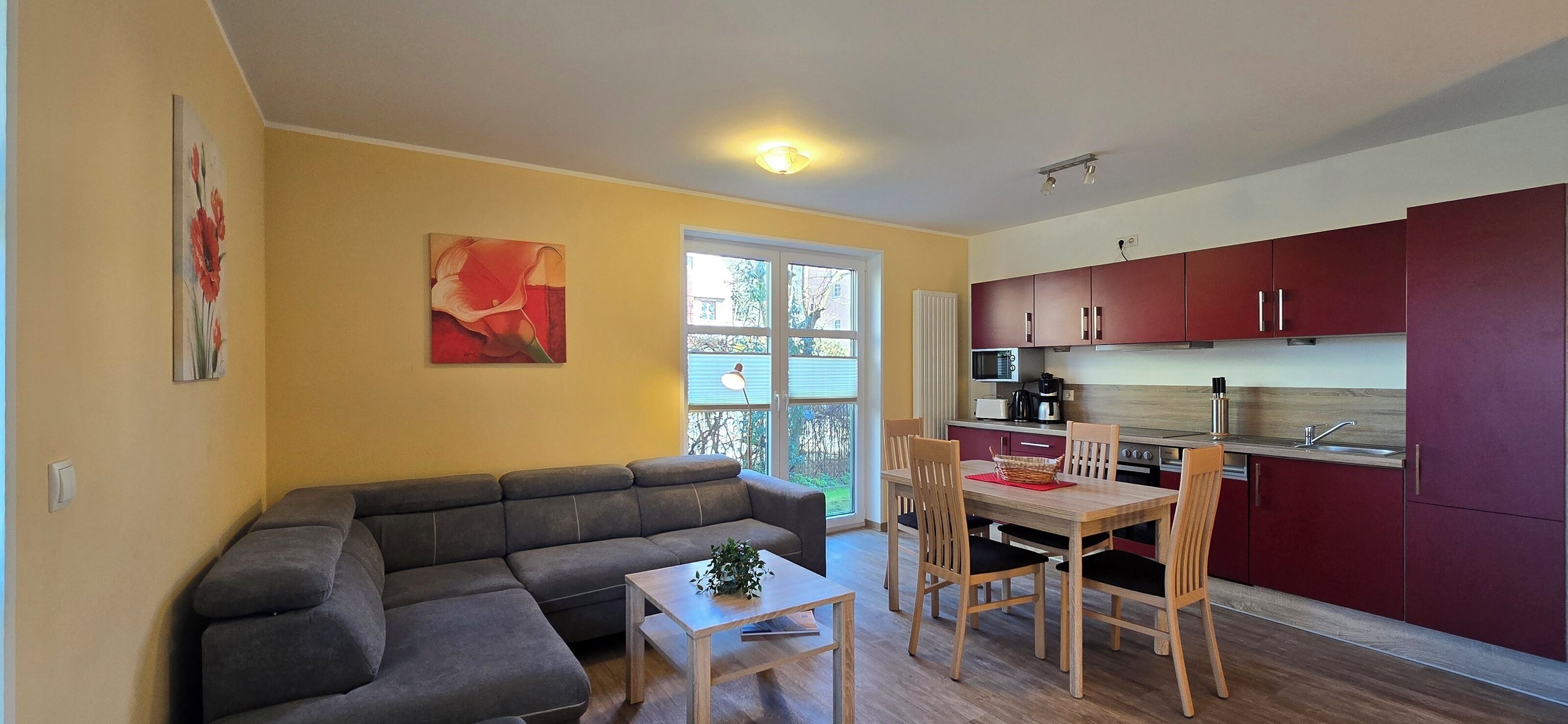 Ein modernes Wohnzimmer mit einem grauen Ecksofa, einem Holzessttisch mit vier Stühlen und einem kleinen Beistelltisch. Ein abstraktes, rotes Gemälde hängt an der Wand.