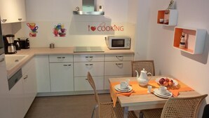 Una cocina compacta con armarios blancos, microondas, hervidor de agua y cafetera. Hay un adorno de pared con temática culinaria y un estante pequeño con objetos decorativos.