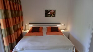 Una cama con ropa de cama blanca y naranja, dos mesitas de noche con lámparas, un cuadro en la pared y una alfombra a rayas en el suelo.