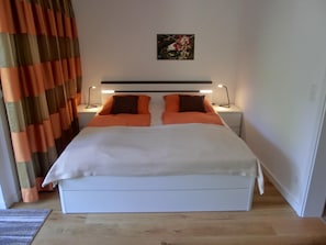 Un lit avec une literie blanche et orange, deux tables de chevet avec des lampes, un tableau au mur et un tapis rayé sur le sol.