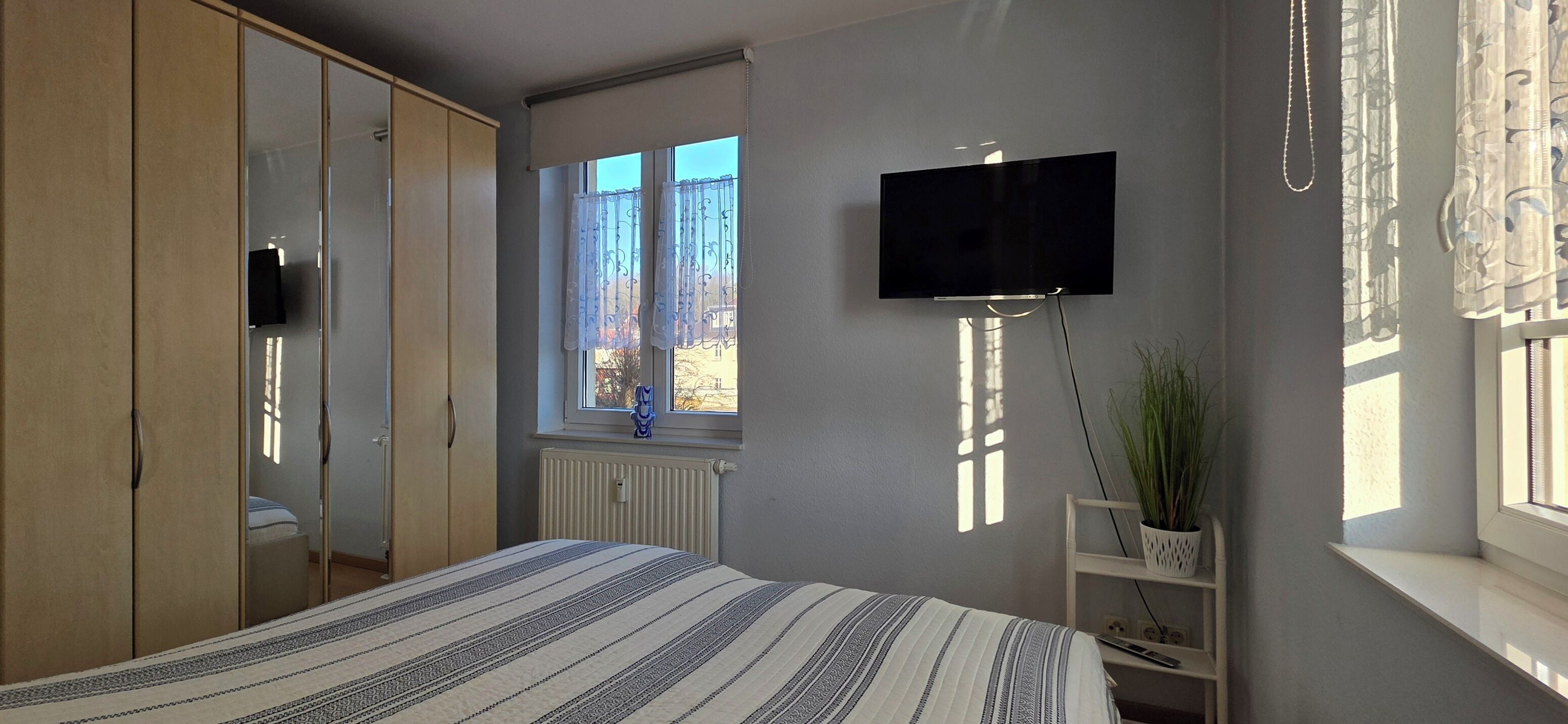Un dormitorio con cama, televisor montado en la pared, ventana con cortinas, radiador y una planta sobre una estantería.