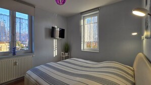 Een slaapkamer met een bed, twee ramen, een televisie en een radiator.