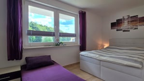 Um quarto com cama, janela com cortinas, mesa de cabeceira com abajur e uma obra de arte na parede.