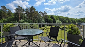Uma varanda com uma mesa redonda e quatro cadeiras, rodeada por um guarda-corpo de metal, com vista para um jardim bem cuidado e uma área arborizada.