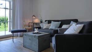 A modern living room with a dark grey sofa, a small wooden table, a lamp, and a view of greenery outside. - Strandnahe 3-zimmer-ferienwohnung für 4 Pers. mit Terrasse (Graal-Müritz)