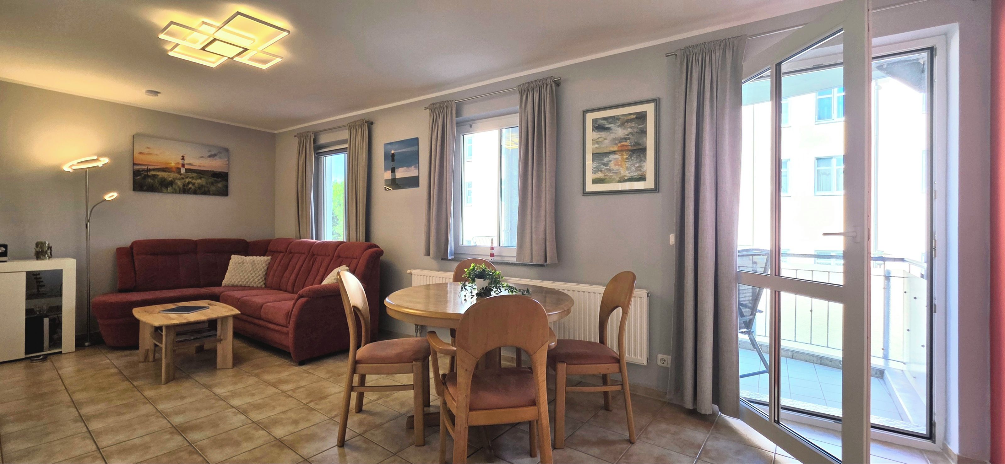 Ein Wohnzimmer mit einer Couch, einem kleinen Tisch und einem Essbereich mit Stühlen. Es gibt Fenster mit Vorhängen, eine Heizung und ein Bild an der Wand.