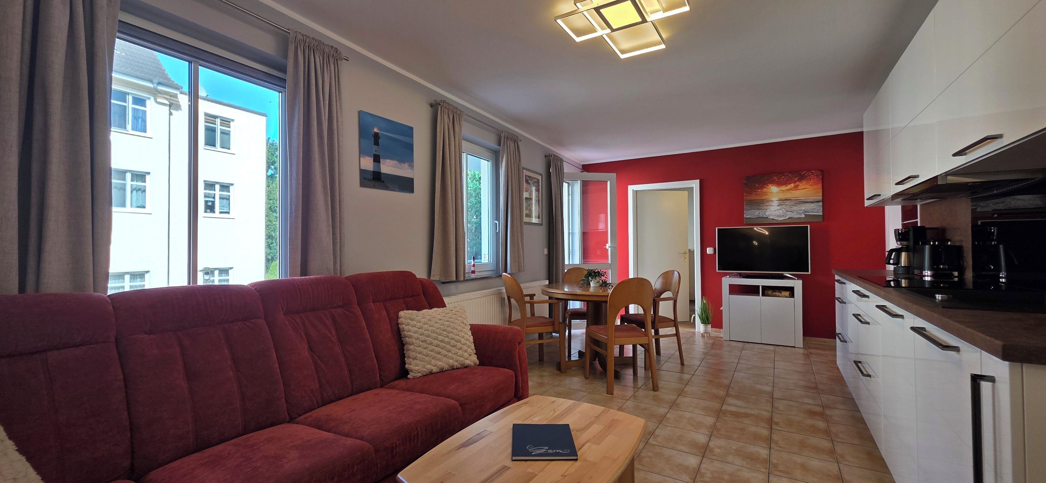 Un salon avec un canapé rouge, une table basse en bois et un téléviseur. On y trouve également un coin repas avec une table et des chaises, ainsi qu’une cuisine équipée de placards et d’une machine à café.