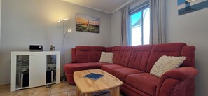 A red sofa with two cushions, a wooden coffee table, a white cabinet with glass doors, and a painting of a lighthouse on the wall. - Strandnahe 2-zi.-fewo mit Balkon für 3 Personen in Ruhiger Lage (Graal-Müritz)