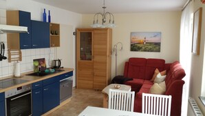 A compact kitchen with modern appliances, a dining area with a red sofa, and a painting of a lighthouse on the wall. - Strandnahe 2-zi.-fewo mit Balkon für 3 Personen in Ruhiger Lage (Graal-Müritz)