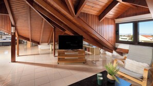 Une pièce spacieuse avec un plafond haut, des poutres apparentes, un téléviseur posé sur un meuble en bois, un canapé et une vue dégagée sur l’extérieur grâce à de grandes fenêtres.