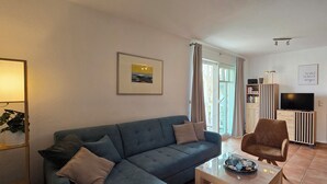 A modern living room with a sofa, armchair, and coffee table. There is a TV, bookshelf, and a door leading to a bedroom. - Strandnahe 2-zi-wohnung im Obergeschoss mit Zwei Balkonen (Graal-Müritz)