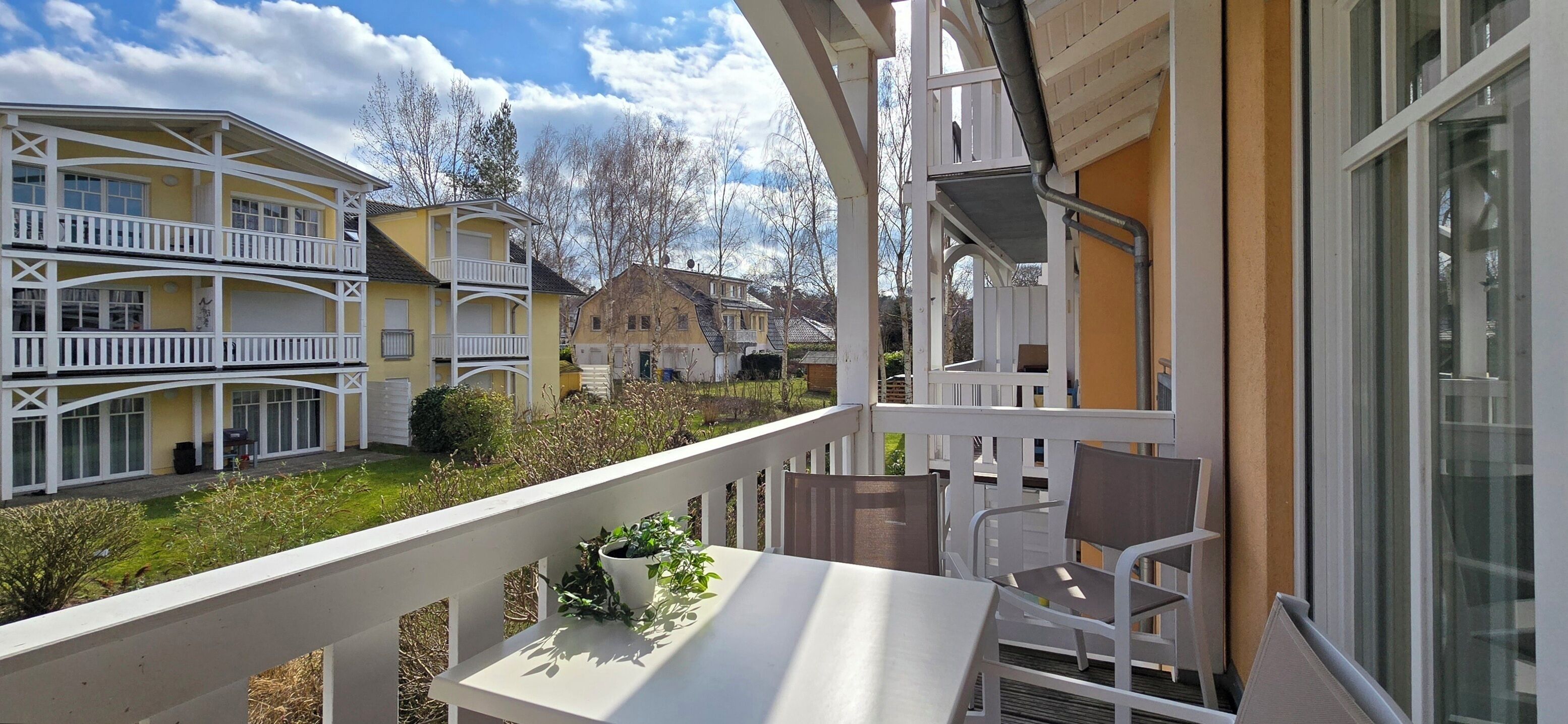 A balcony with a table and chairs, overlooking a courtyard with a yellow building and a stone house.