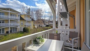 A balcony with a table and chairs, overlooking a courtyard with a yellow building and a stone house. - Strandnahe 2-zi-wohnung im Obergeschoss mit Zwei Balkonen (Graal-Müritz)