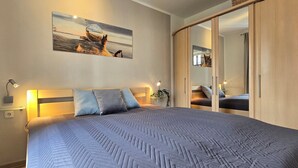 A neatly made bed with a patterned bedspread, a headboard with wall-mounted lights, and a large mirror cabinet with a reflection of a dog. - Strandnahe 2-zi-wohnung im Obergeschoss mit Zwei Balkonen (Graal-Müritz)