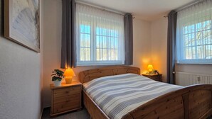 Ein Schlafzimmer mit einem hölzernen Bett, zwei Nachttischen mit Lampen, einem Heizkörper und einem Fenster mit Vorhängen.