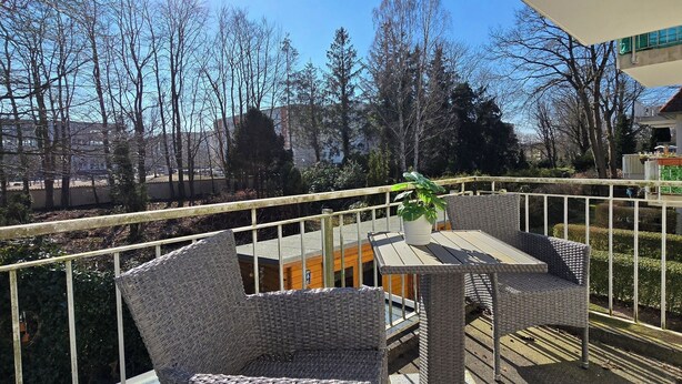 Ein Balkon mit Korbmöbeln, einer Topfpflanze und Blick auf Bäume und Gebäude.