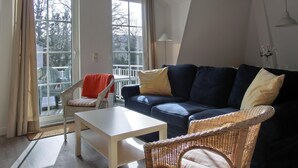 Ein Wohnzimmer mit einer Couch, einem Tisch und einem Korbstuhl. Große Fenster und Blick auf Bäume draußen.